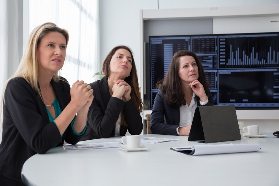 Three women in a business meeting room discussing charts and data on screens.